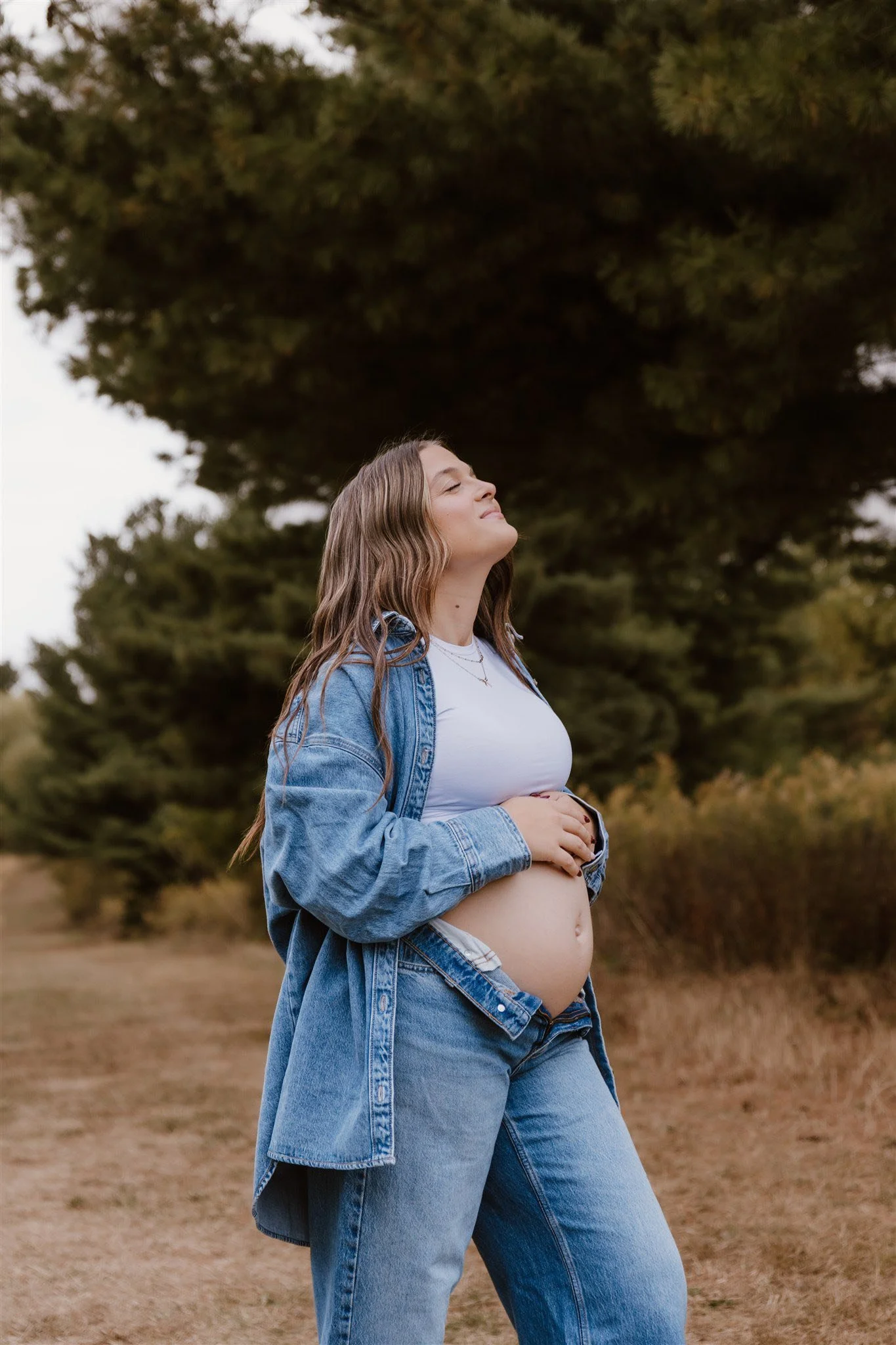 A pregnant woman with long wavy hair, wearing a white crop top, blue jeans, and an unbuttoned denim jacket, standing outdoors with her eyes closed and a content expression on her face, surrounded by trees and grass.