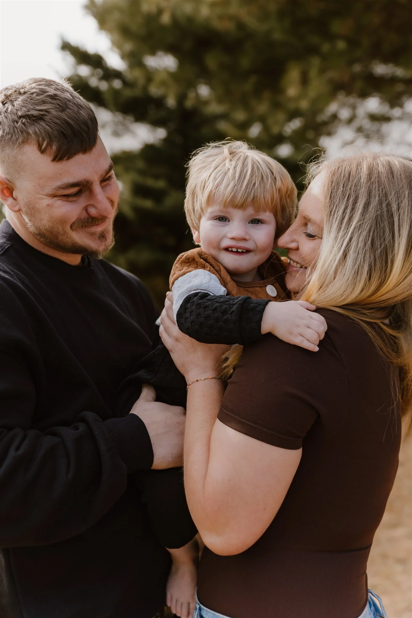 A family of three, a man, a woman, and a young boy, are outside. The woman is holding the boy, and they are all smiling and looking at each other. The background features trees and an outdoor setting.