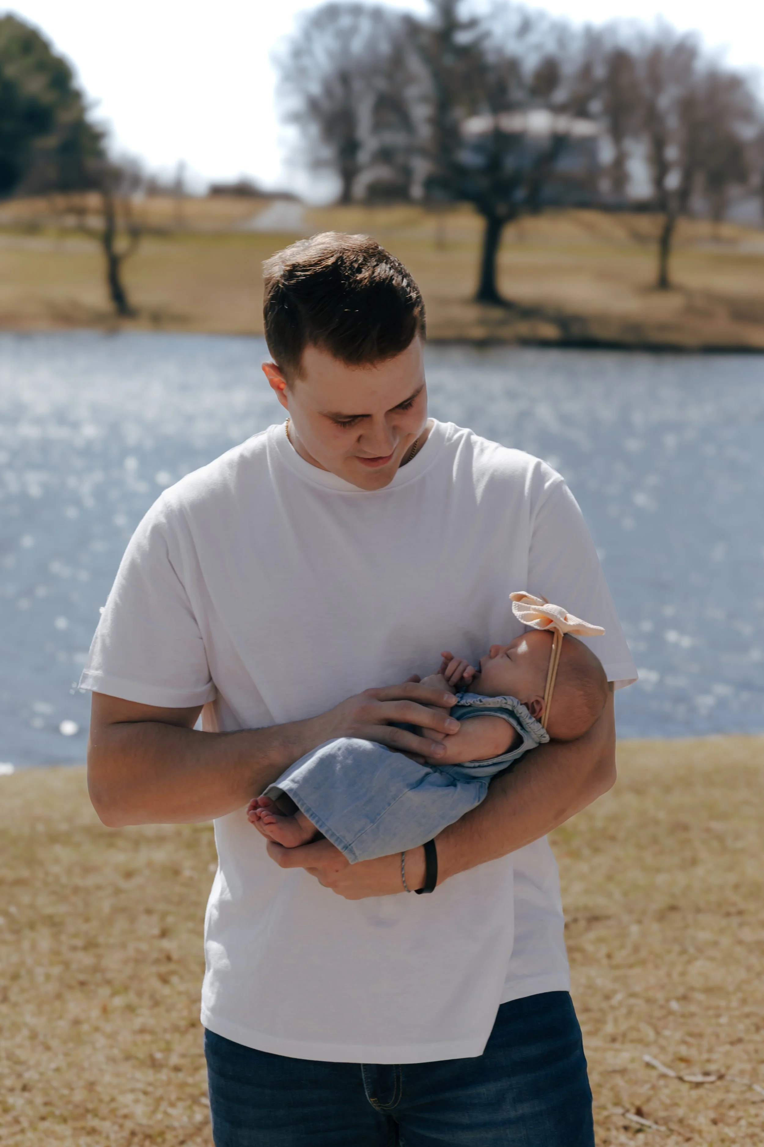 Father holding newborn baby with lake sparkling in the background during Central Illinois family photography session