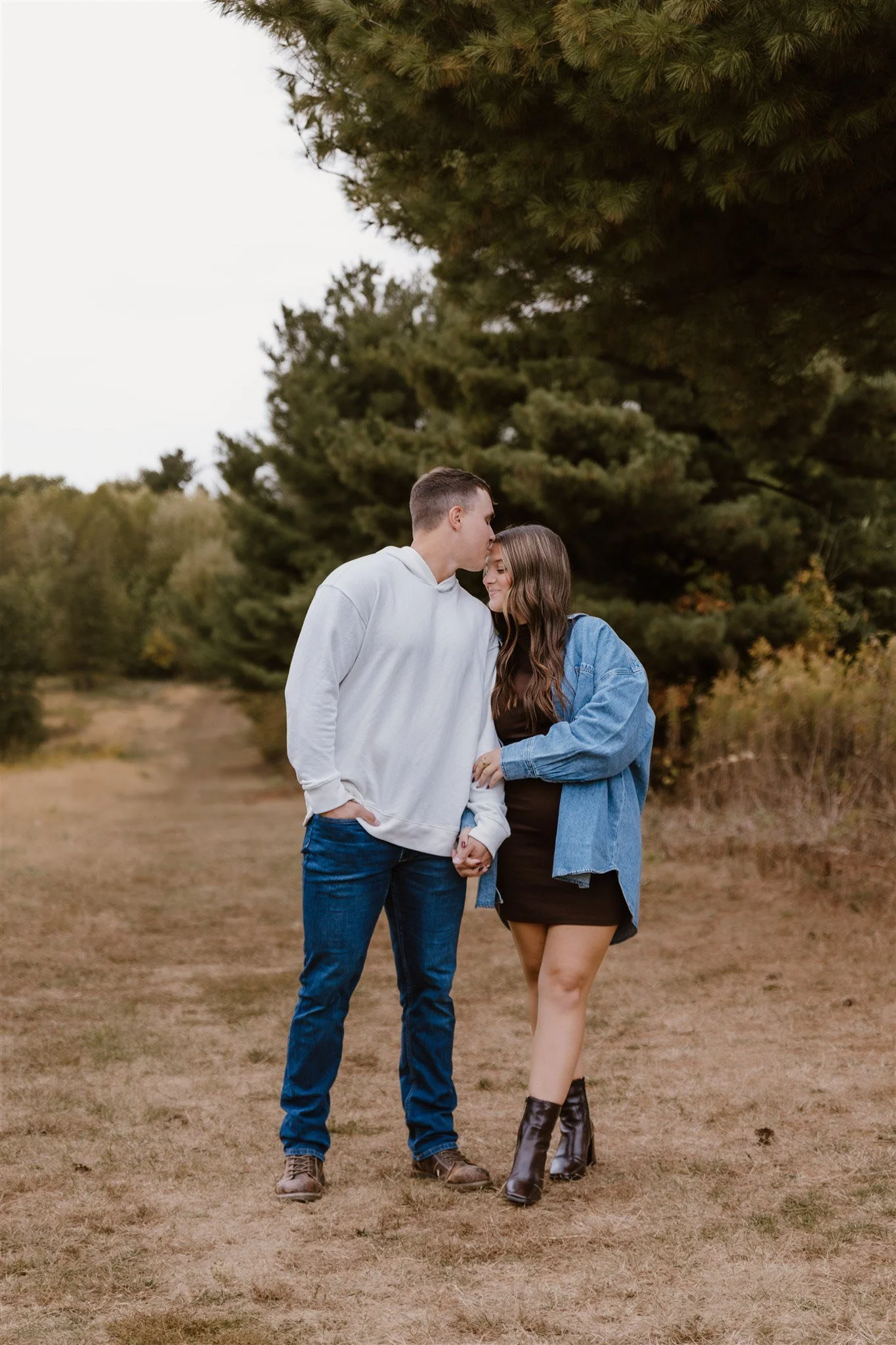 A young couple is standing on a grassy pathway outdoors, holding hands. The man is kissing the woman's forehead, and they are smiling. The man is wearing a white hoodie and jeans, and the woman is wearing a black dress, a blue denim jacket, and black