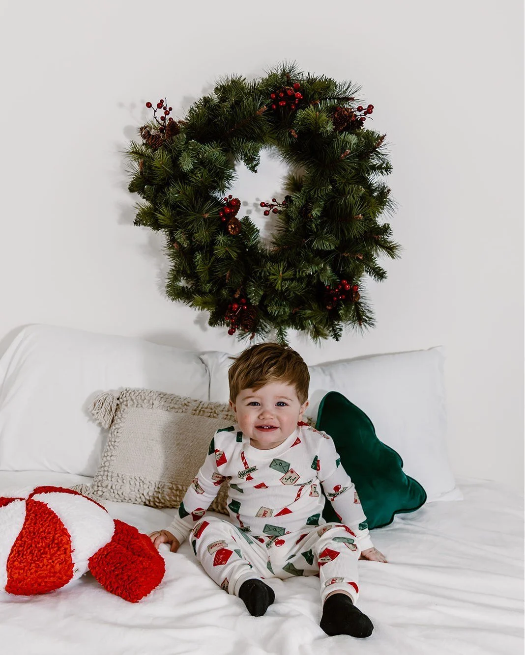 A young boy in holiday pajamas sitting on a white bed with a Christmas stocking. Behind him, a white wall with a holiday wreath made of greenery and red berries.