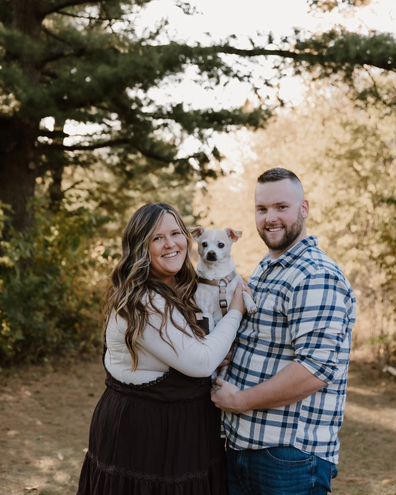 A smiling woman and man holding a small dog outdoors with trees and sunlight in the background.