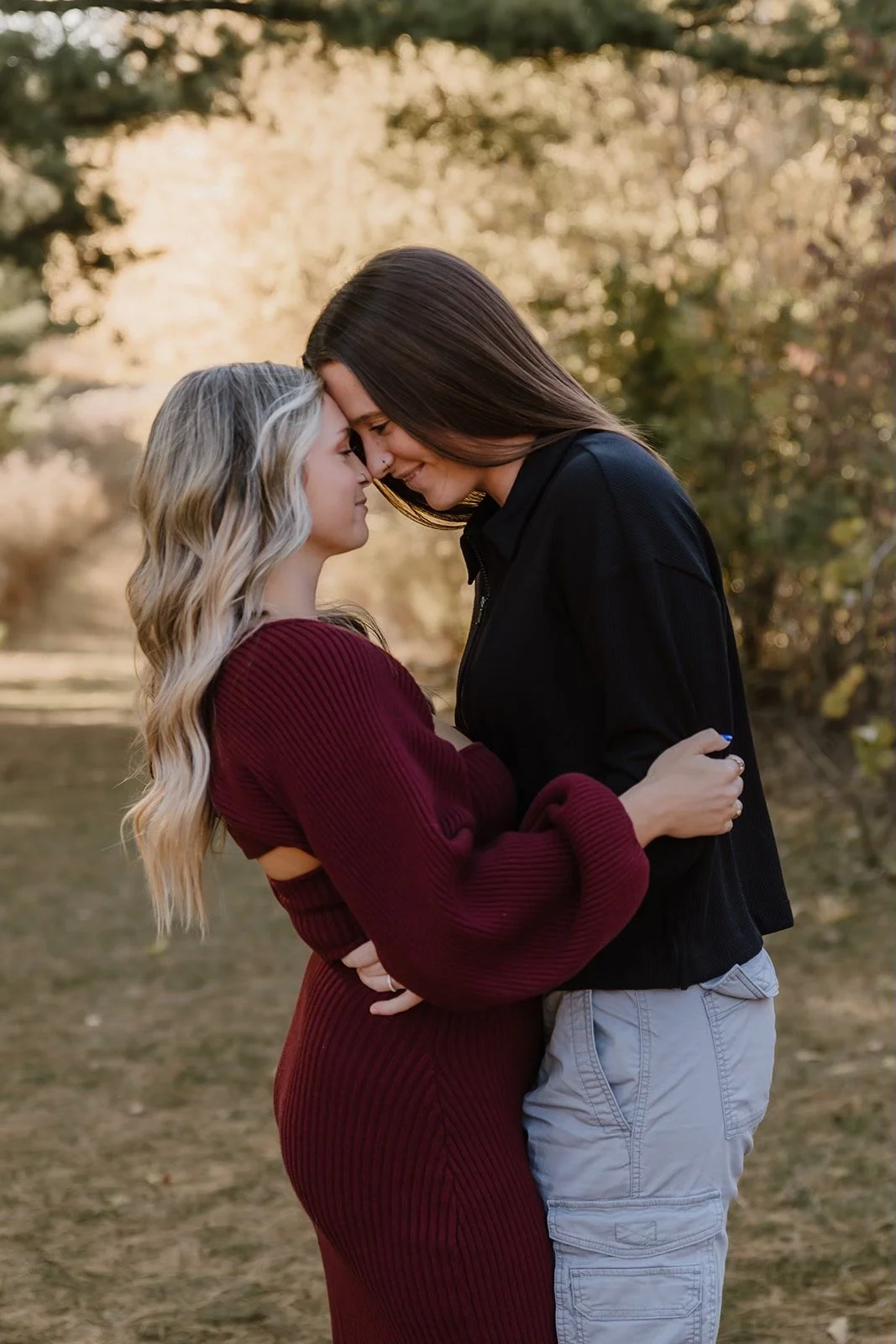Two women standing close together outdoors, touching foreheads, with trees in the background.
