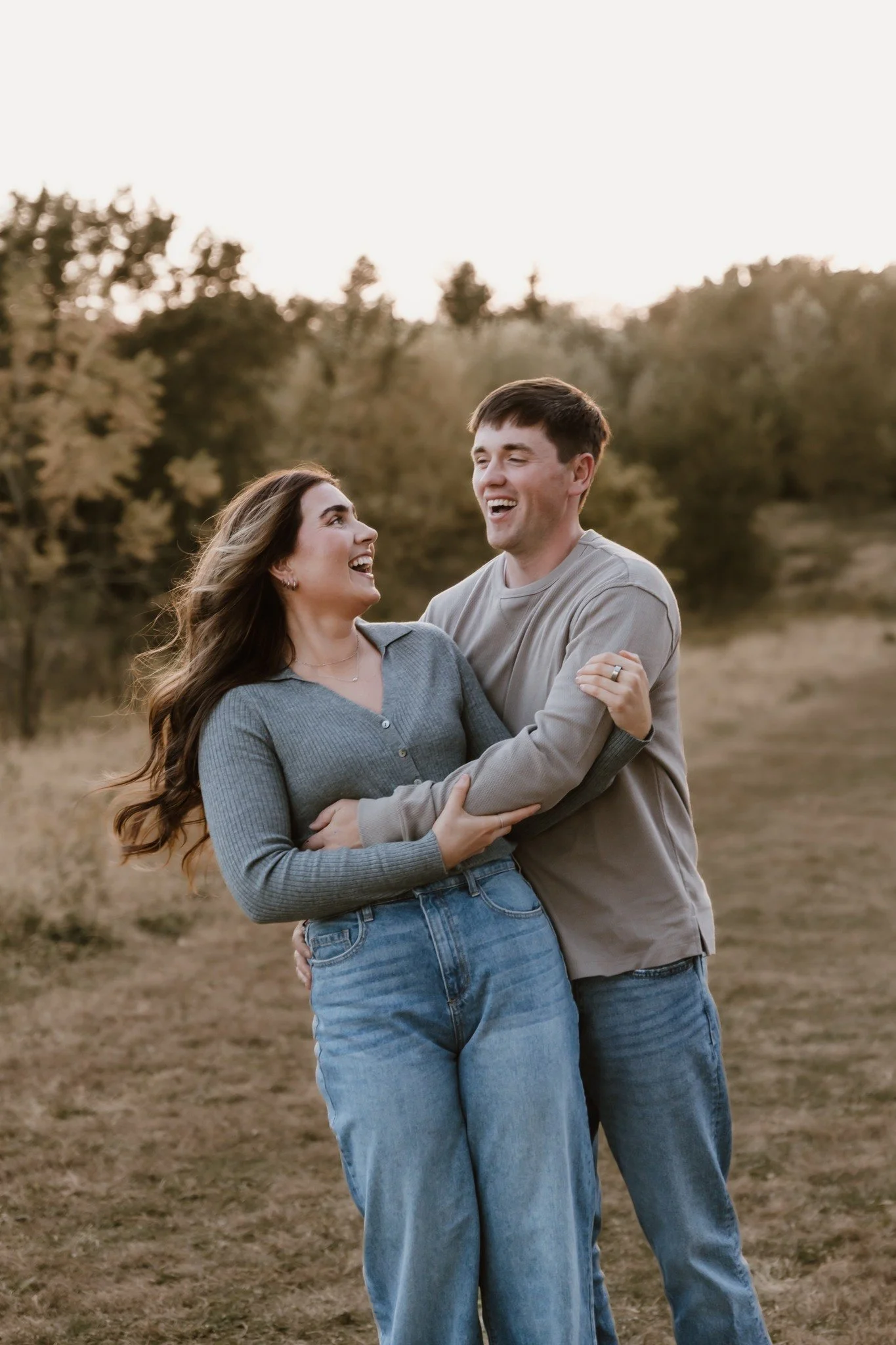 A couple laughing and embracing outdoors in a field with trees in the background, during sunset.