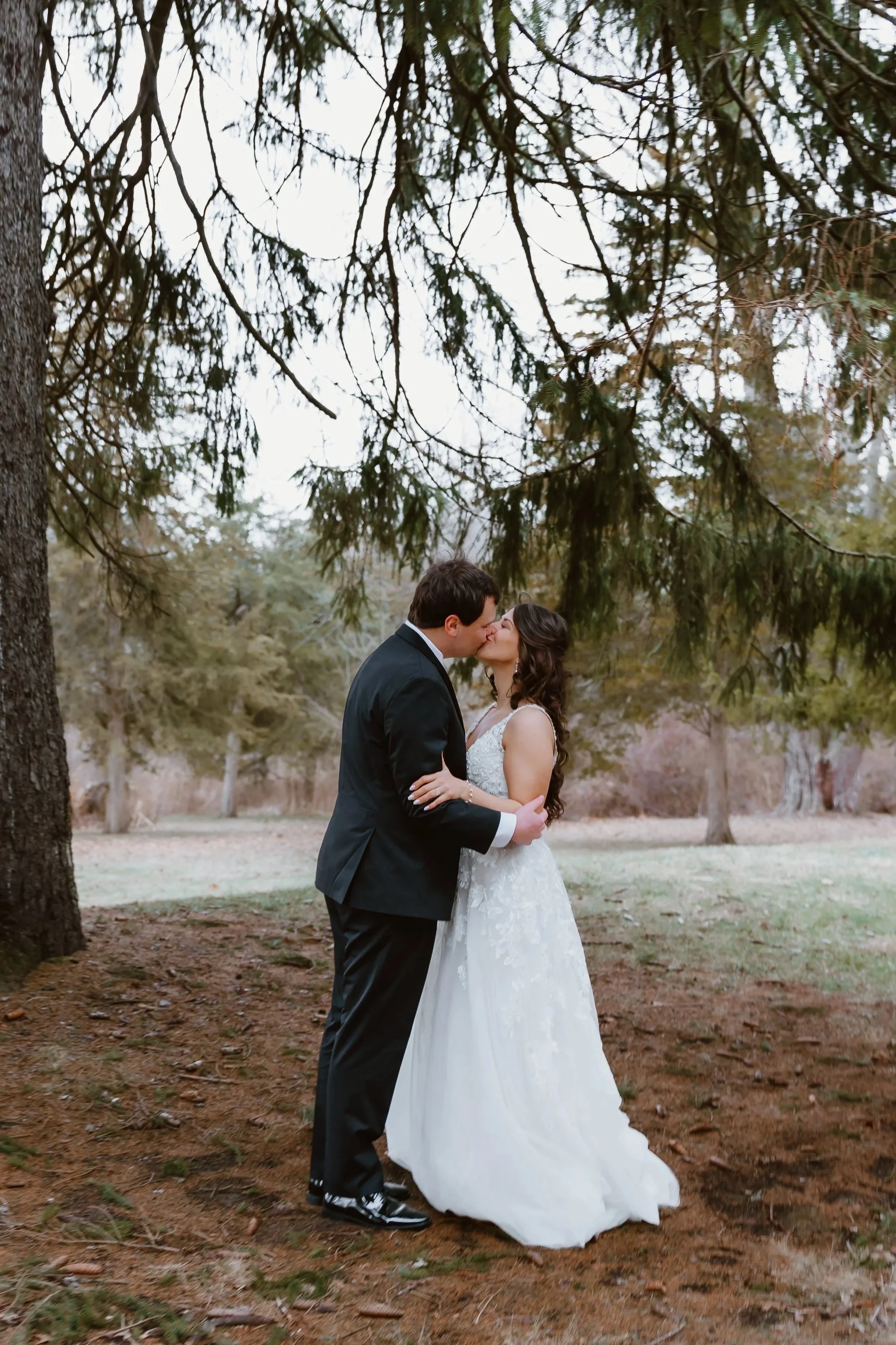 A couple in wedding attire kissing in an outdoor park setting under tree branches.