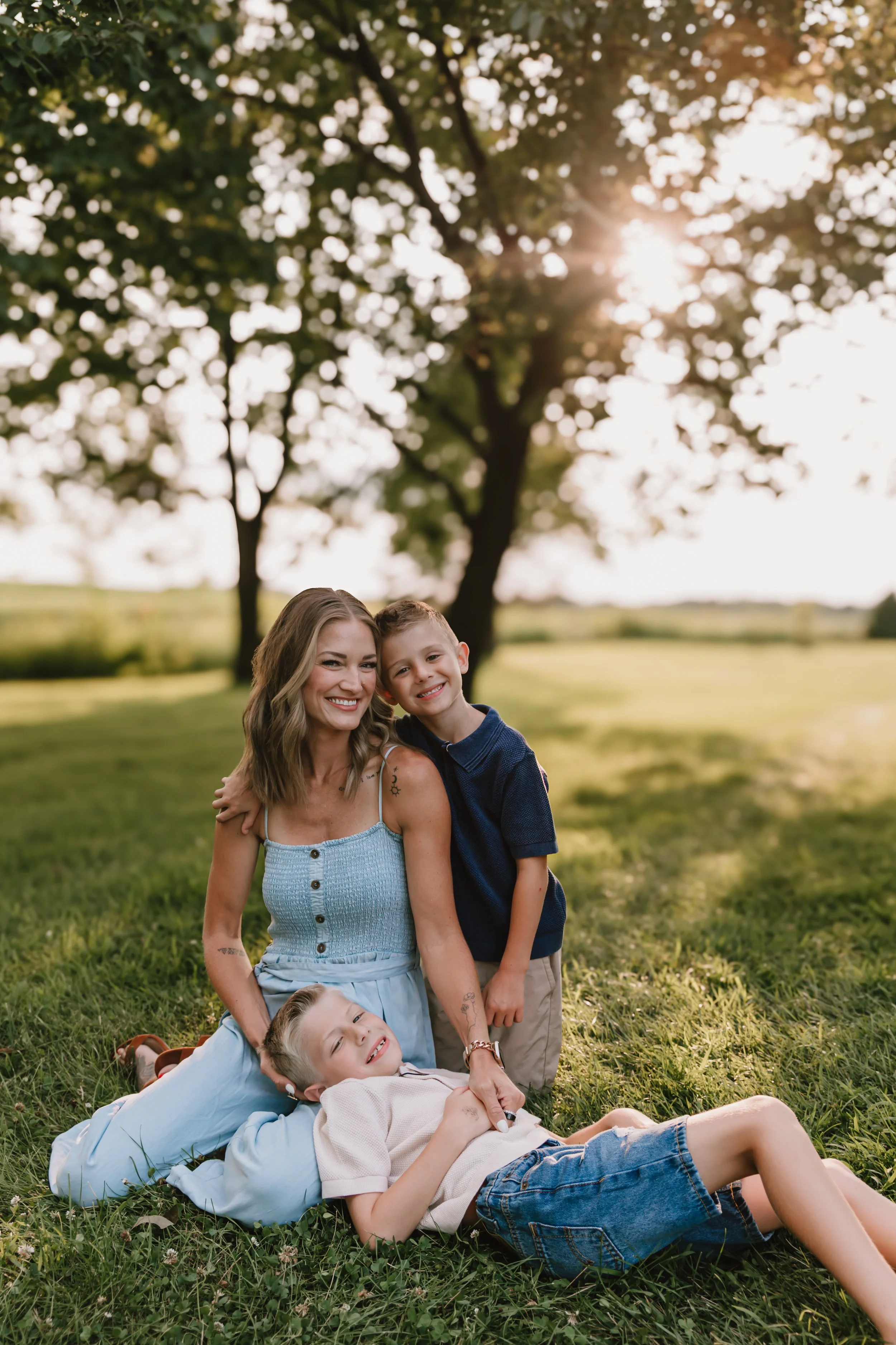 Mother smiling with her two boys beneath large trees during golden hour family photos in Peoria, Illinois.