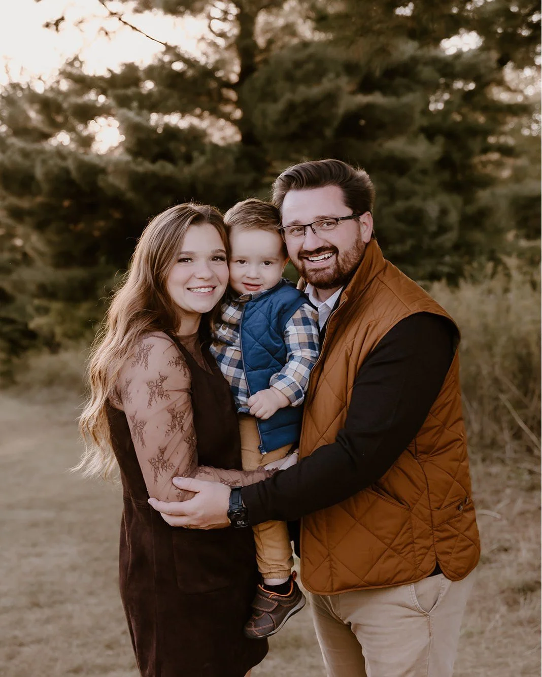 A family of three outdoors at sunset, smiling and hugging, with large trees in the background.