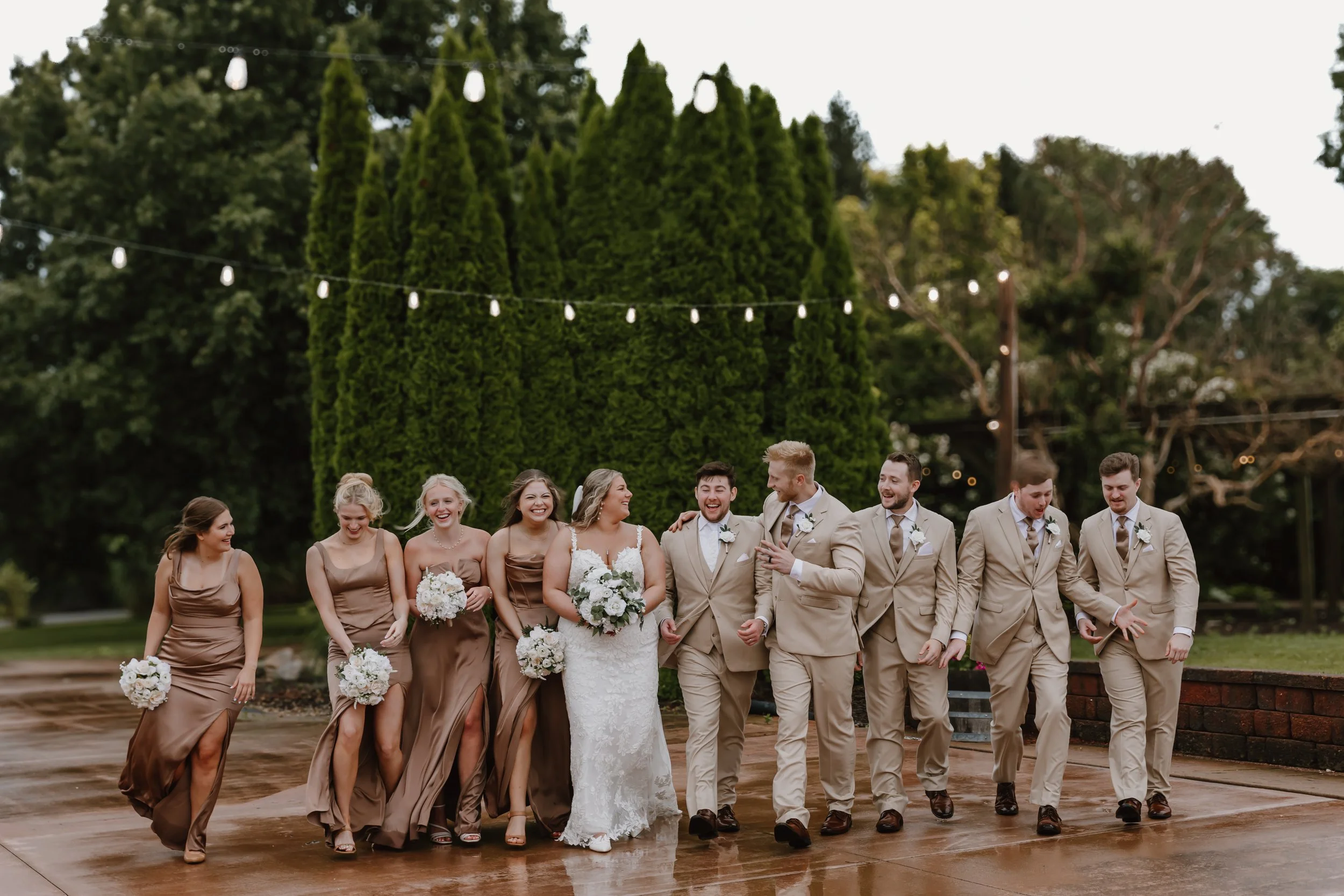 A wedding party walking outdoors on a wet surface, with women in rose-colored dresses and men in beige suits, all holding bouquets, smiling and laughing in Edwards, illinois