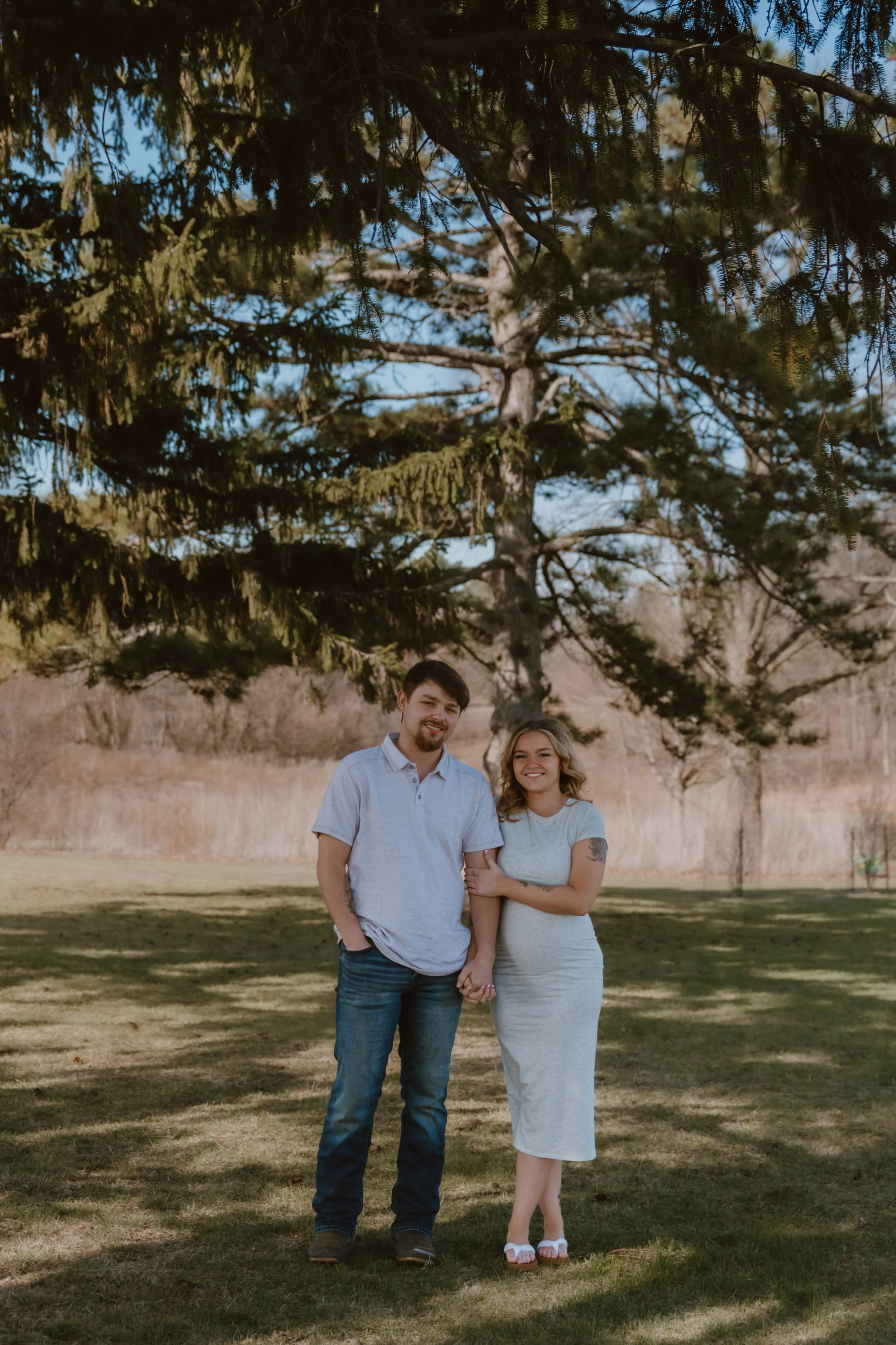couple standing under pine trees during spring maternity session in peoria illinois