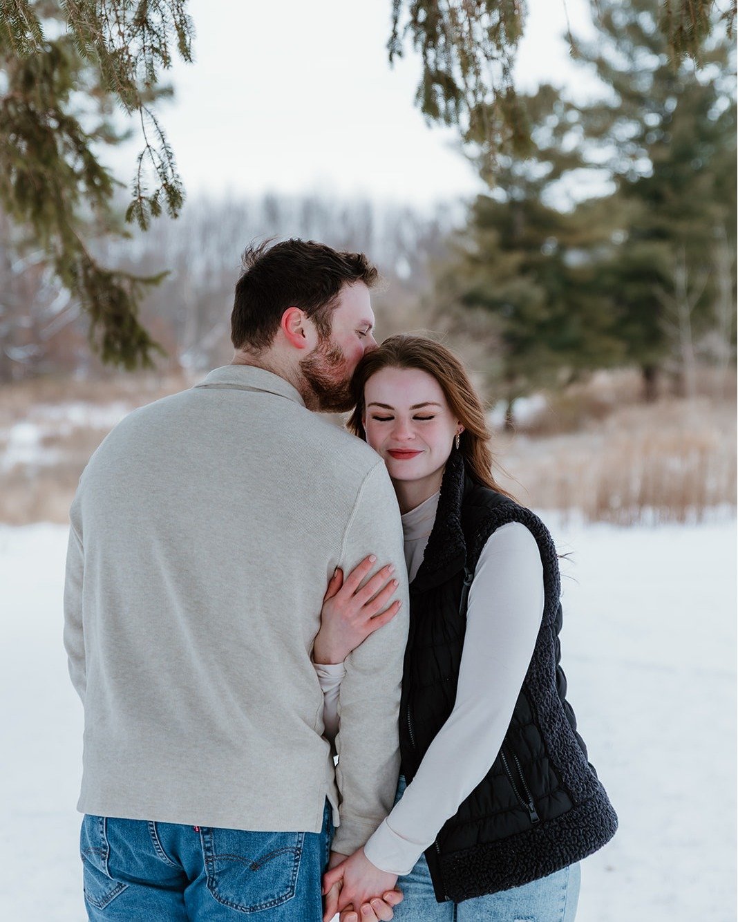 A couple hugging outdoors in a snowy landscape with trees in the background, the man kissing the woman's head.