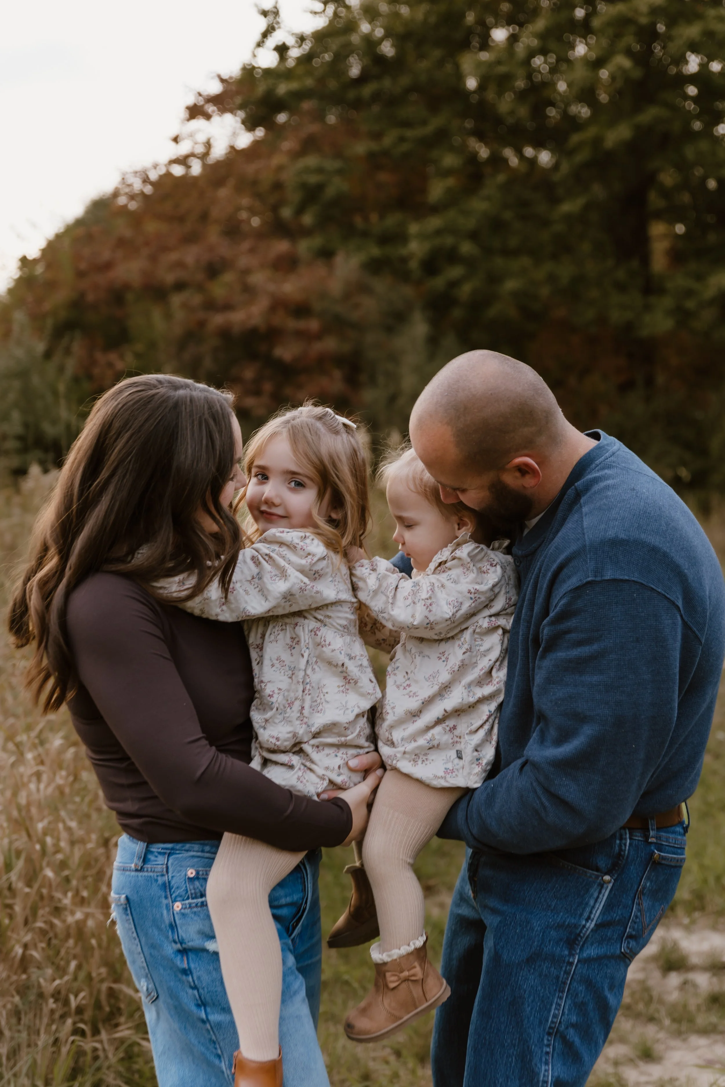 A family of four enjoying outdoors during autumn. Parent woman with long brown hair, parent man with a shaved head, and two young girls in matching outfits. They are sitting in a grassy field with trees in fall colors in the background.