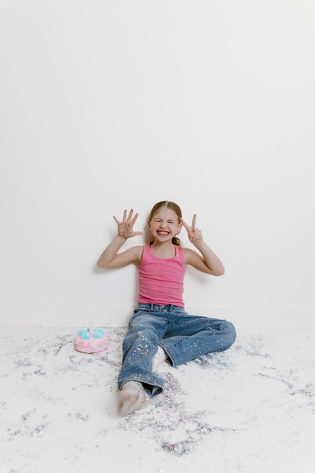 A young girl in a pink tank top and jeans sitting on the floor covered in white powder, smiling and making peace signs, with a small pink and blue birthday cake nearby.