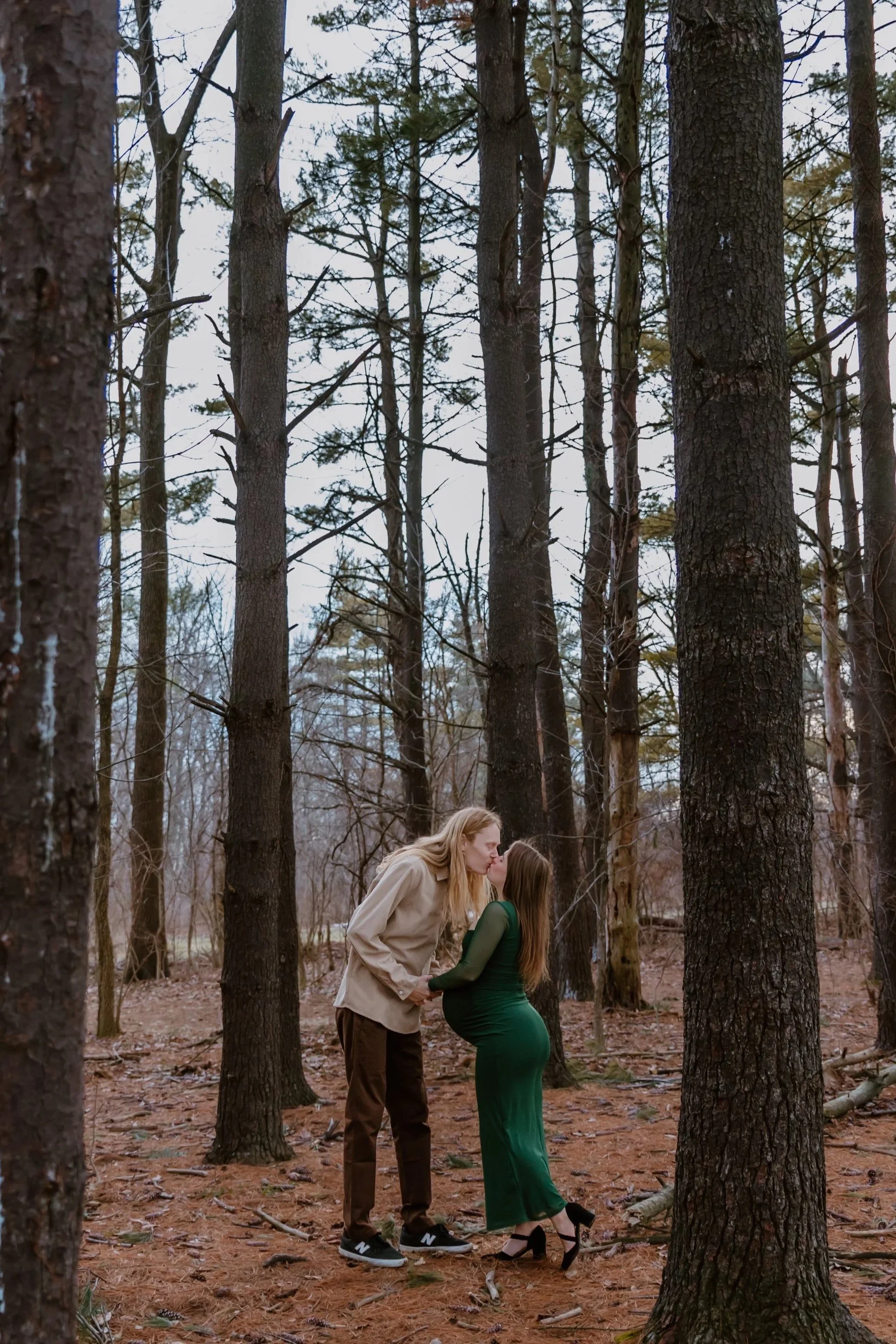 A woman in a green dress and heels kissing a pregnant woman in a beige jacket and dark pants in a forest with tall trees and brown pine needles.