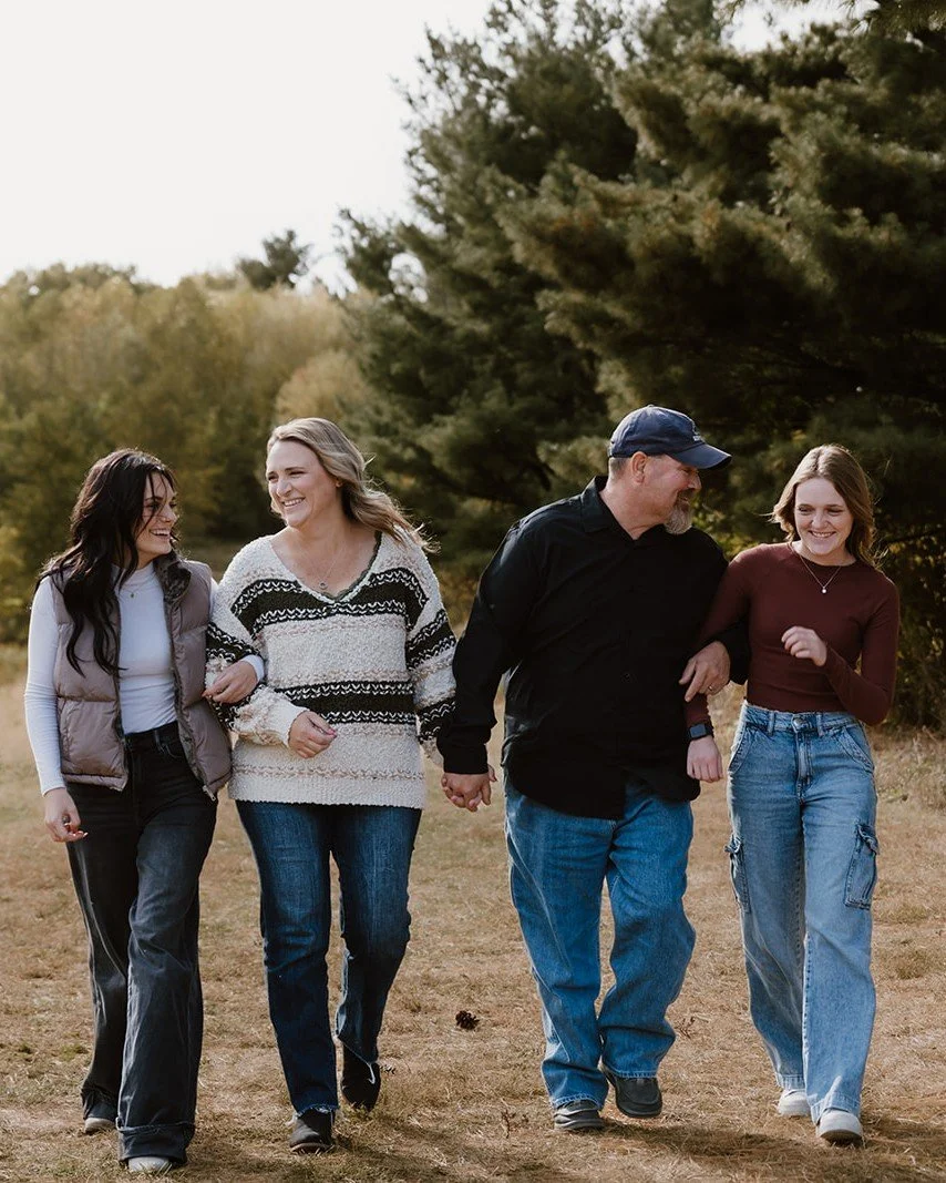 A family of four walking outdoors in a grassy park on a clear day. They are holding hands and smiling, with trees in the background.