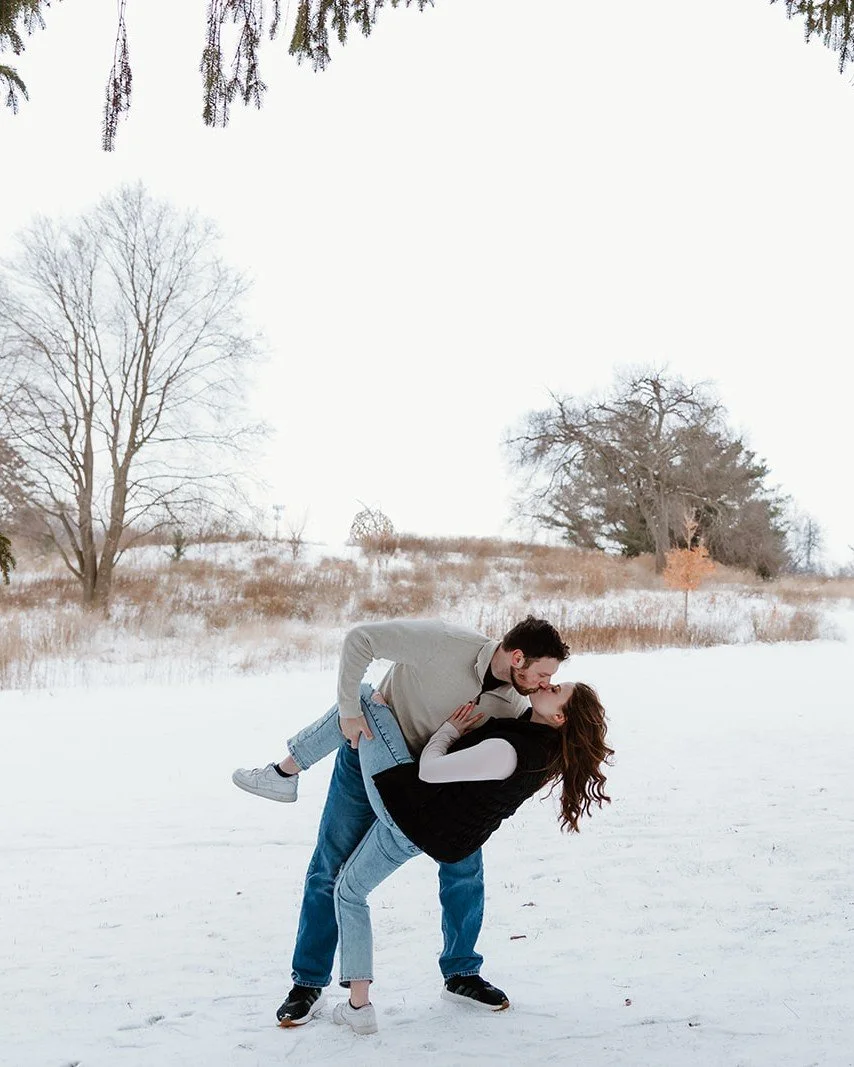 A couple sharing a kiss in a snowy outdoor field, with trees in the background and wintery landscape.