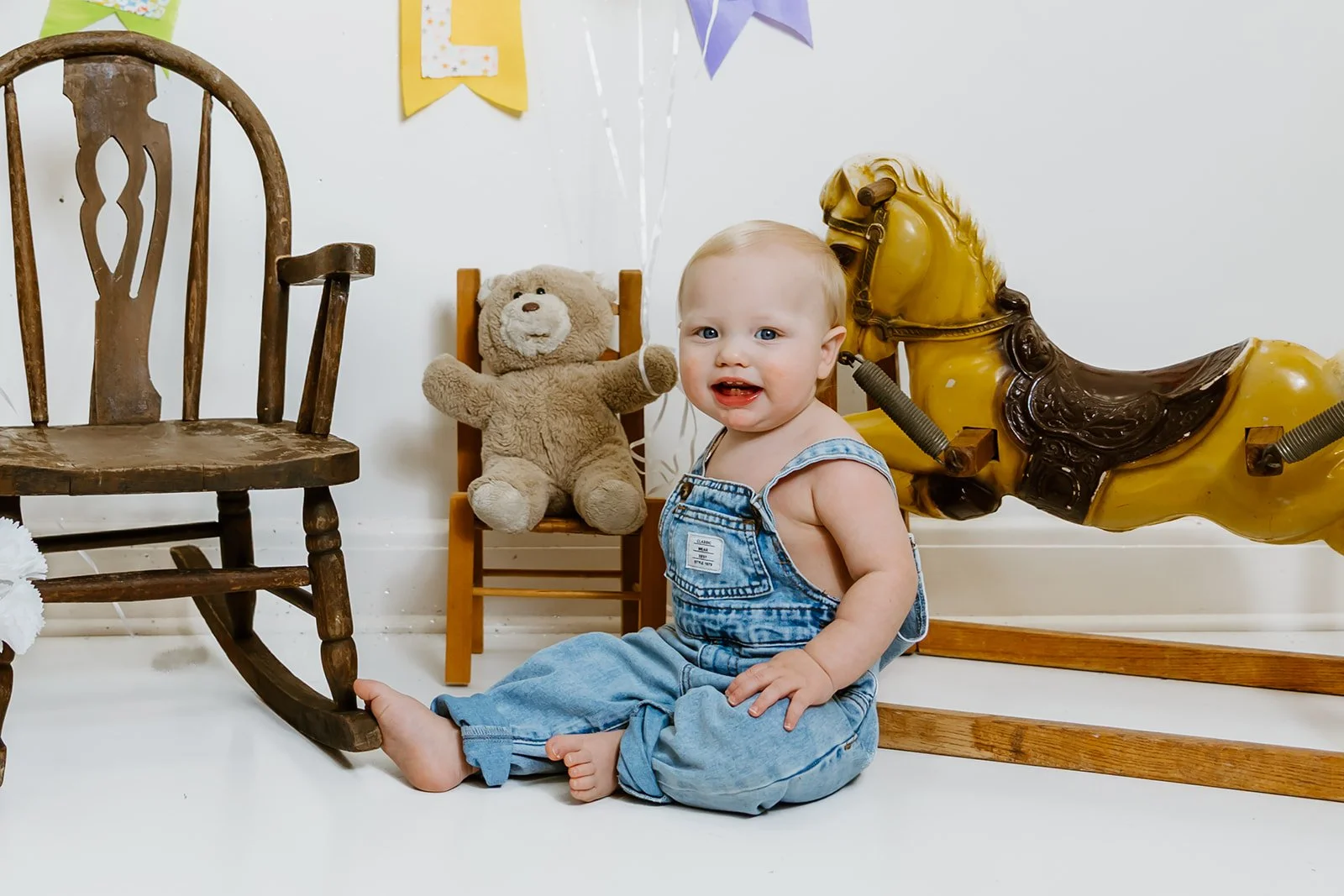 A toddler sitting on the floor next to a vintage wooden rocking horse with a yellow wooden horse in the background, a teddy bear, and a wooden chair, with colorful banners hanging on a white wall.
