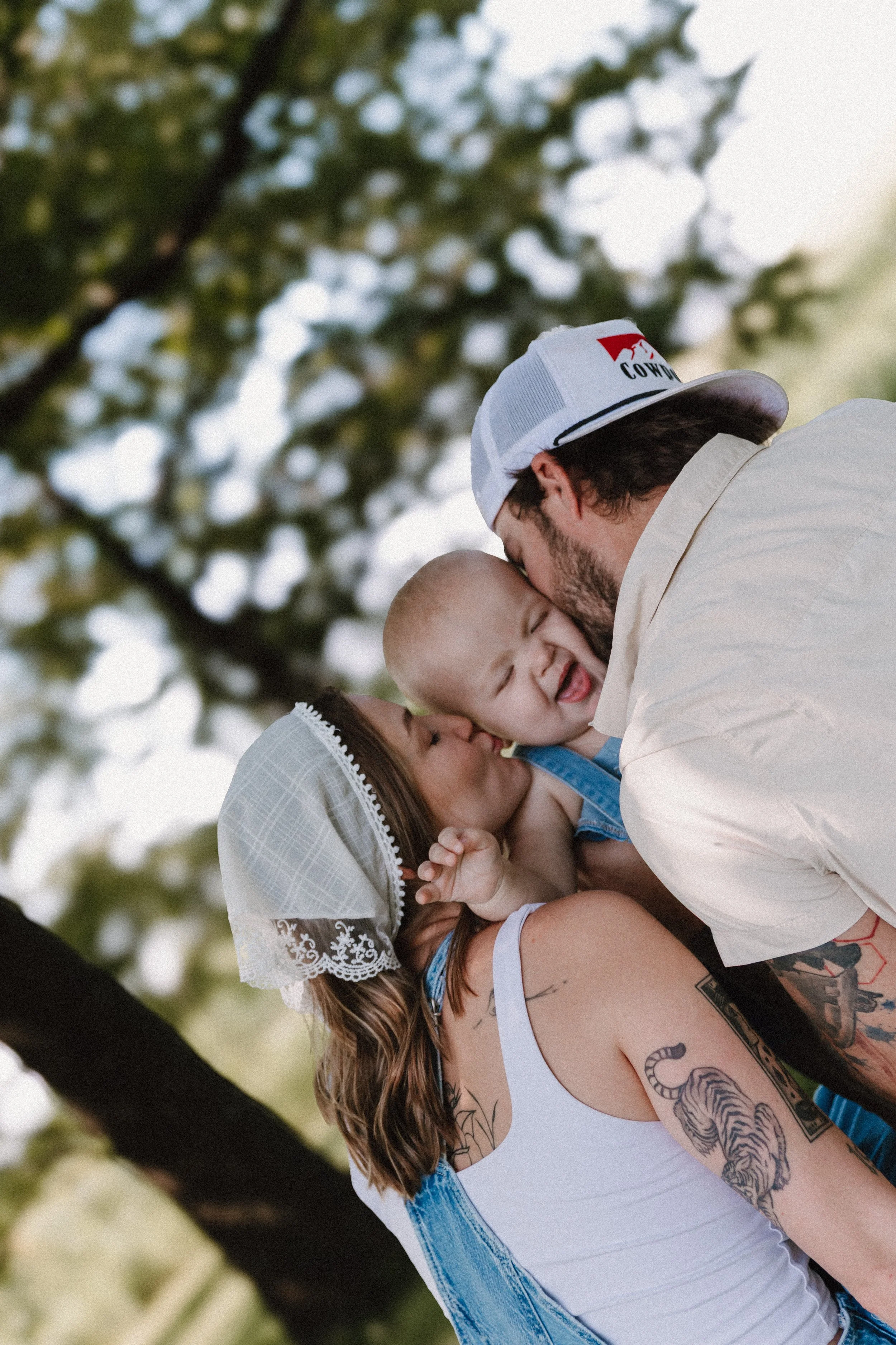 A man with Tattoos holding a child and kissing her face while a woman looks on, outdoors with trees in the background in East Peoria illinois