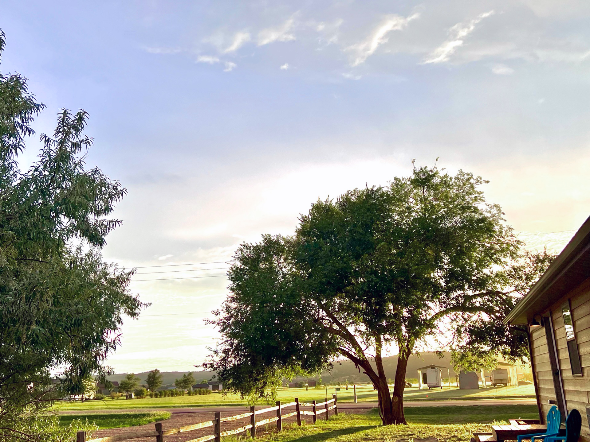 A scenic view of a rural landscape with trees, a wooden fence, and a clear sky at sunset. A building and some structures are visible in the distance on a grassy field.
