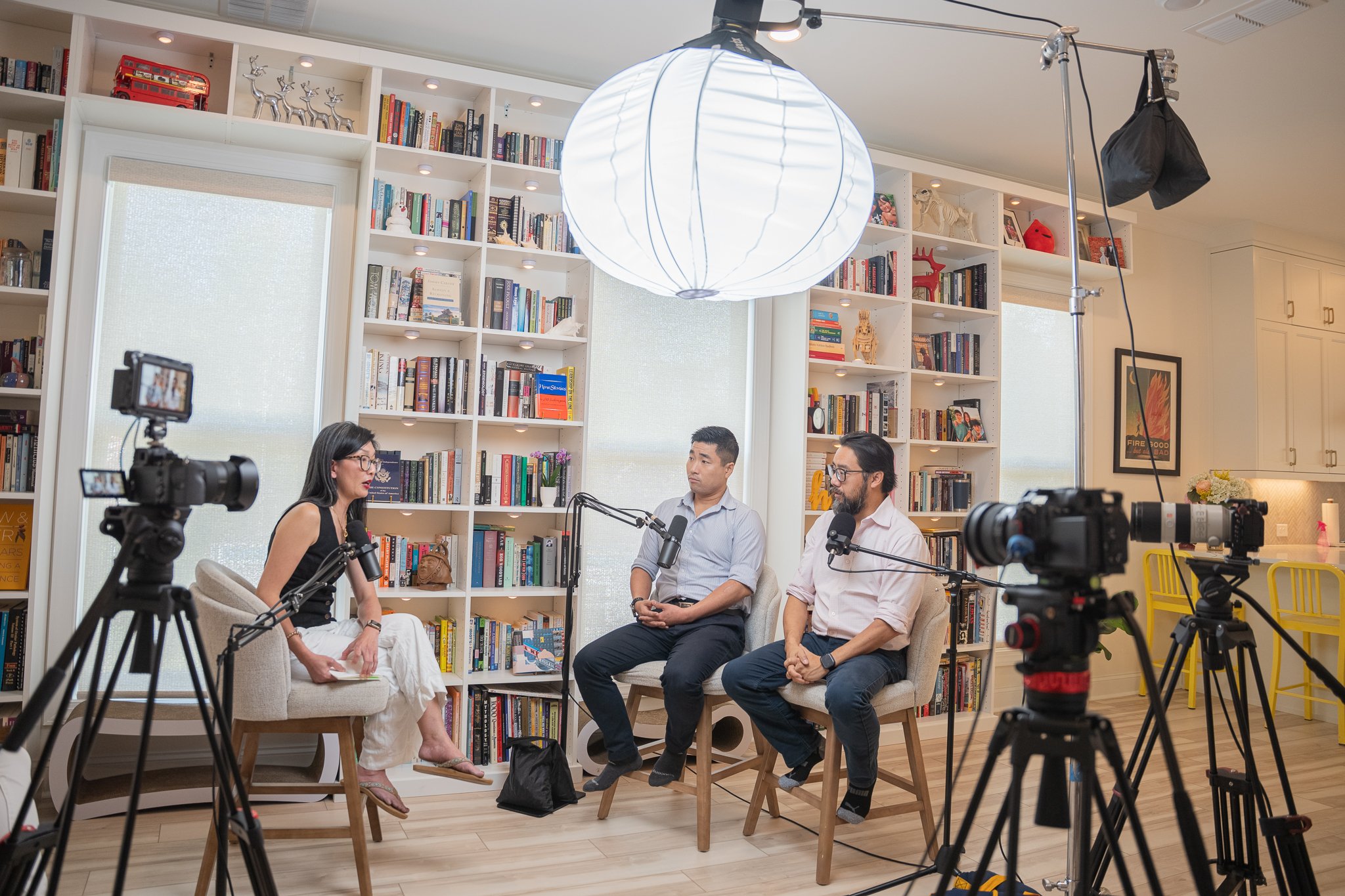 Three people participating in a podcast recording in a cozy, well-lit room with bookshelves, a large ceiling light, and professional video and audio equipment.