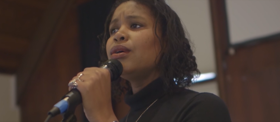 Whitney Parnell, a Black woman with curly hair, singing into a microphone with a dark purple overlay.