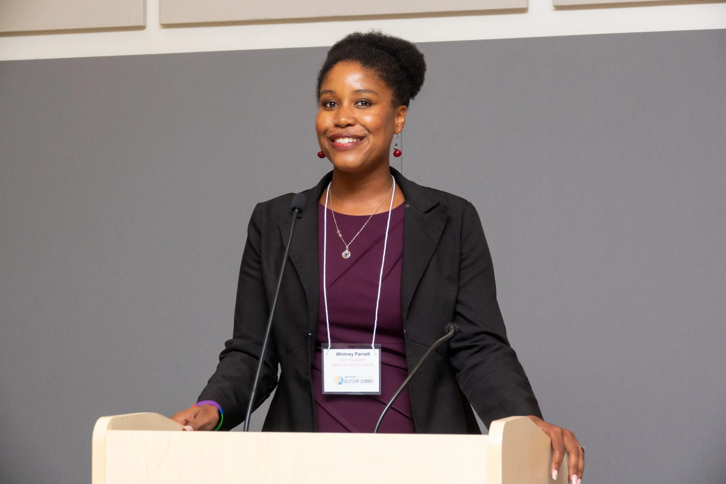 Whitney Parnell speaking behind a podium at a conference, smiling and wearing a black blazer, purple dress, and jewelry.