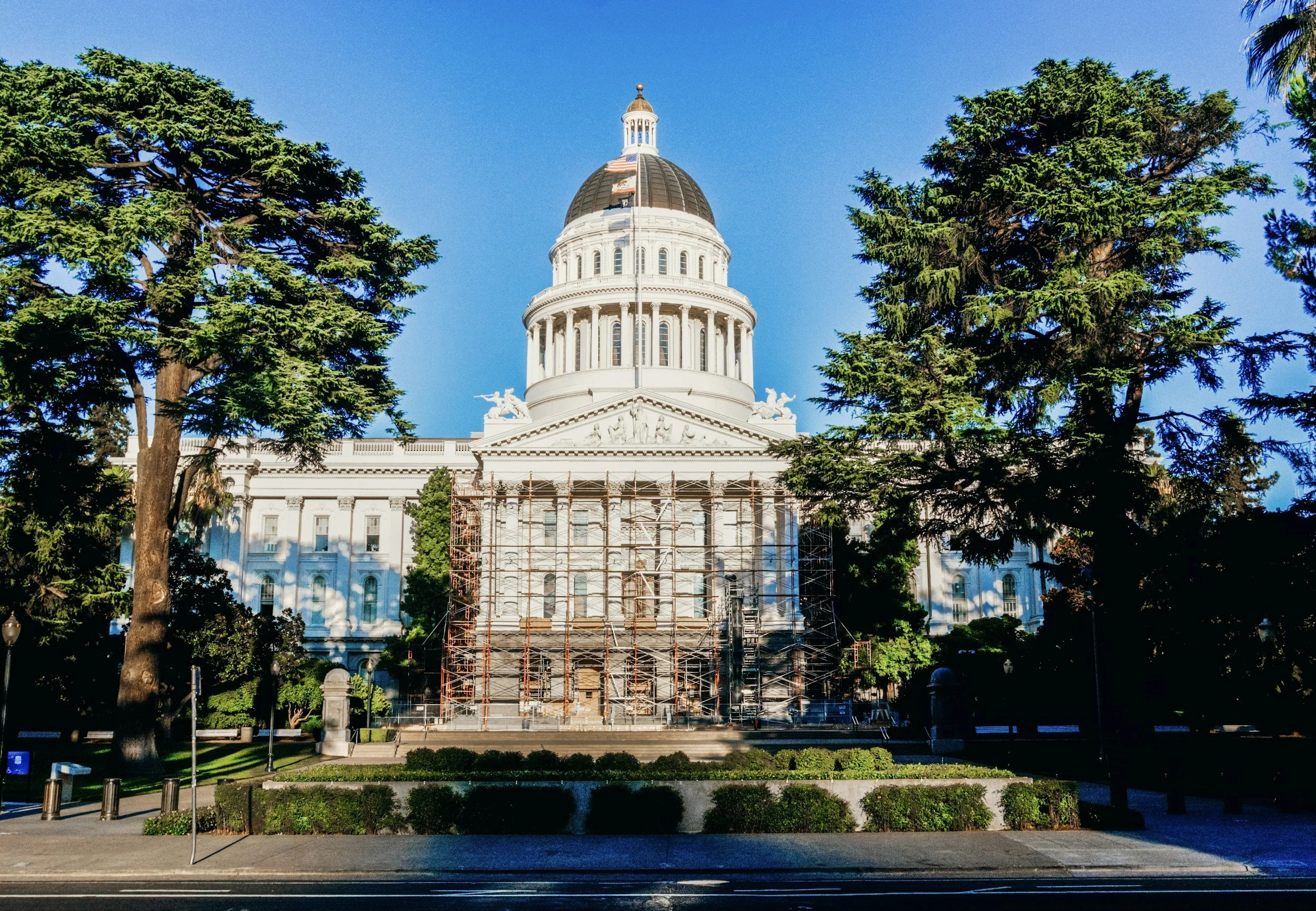 A white, domed State Capitol building with scaffolding on its lower facade, framed by large green trees against a clear blue sky.