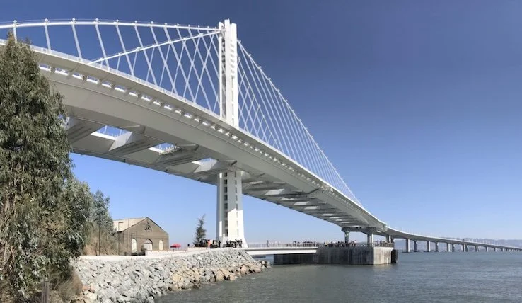 White suspension bridge spanning blue bay beside rocky shoreline and small historic building under clear sky