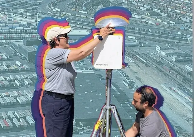 Two people adjust a white device on a tripod overlooking a shipping yard. They have a colorful, multi-layered "echo" effect around them.