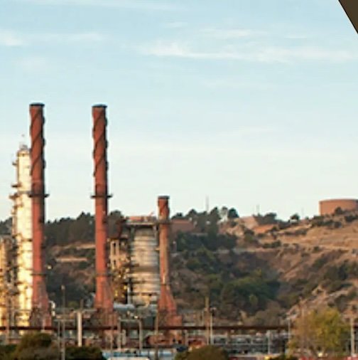 A wide landscape photo of an industrial refinery with two tall, red-and-black smokestacks in the foreground and rolling hills behind.
