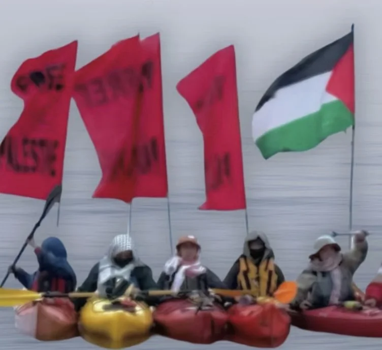 A group of kayakers in colorful boats hold red protest flags and a Palestinian flag while lined up on the water.