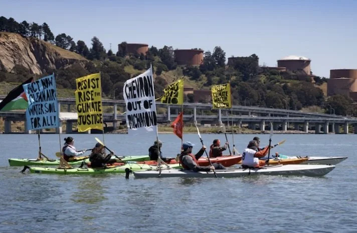 Environmental activists in kayaks carry protest banners on a waterway near a bridge and industrial facility, calling for climate justice.