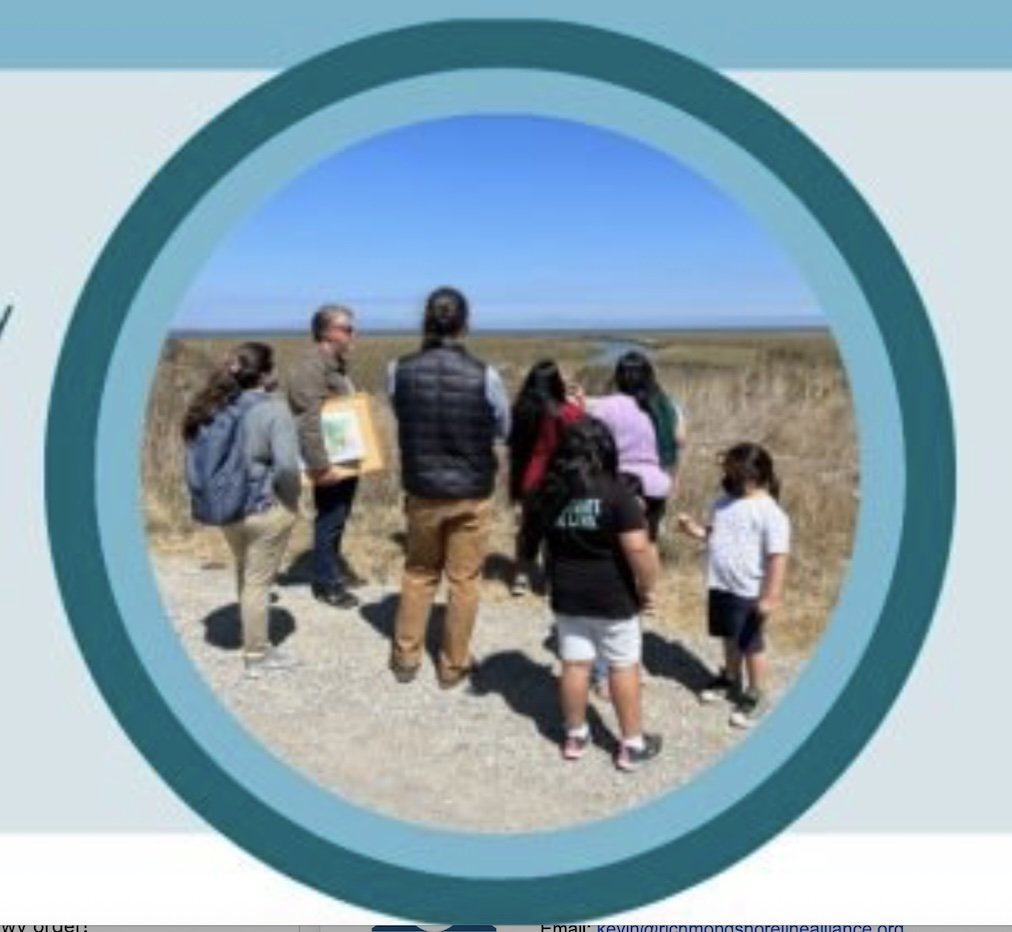 A group of people standing on a gravel path outdoors, looking at a grassy marsh under a clear blue sky. One person holds a poster.