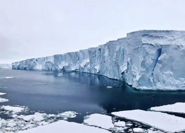 A massive, sheer blue-white ice shelf stretches across dark Antarctic waters under a pale sky, with jagged ice chunks floating in the foreground.