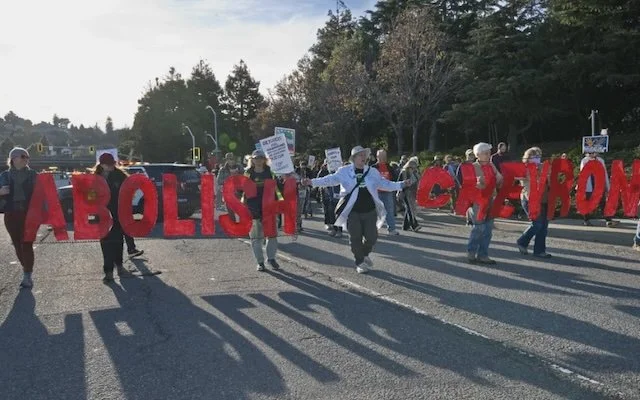 Protesters march across a street holding a large, red sign that reads "ABOLISH CHEVRON" in bold letters. People hold smaller signs behind it.