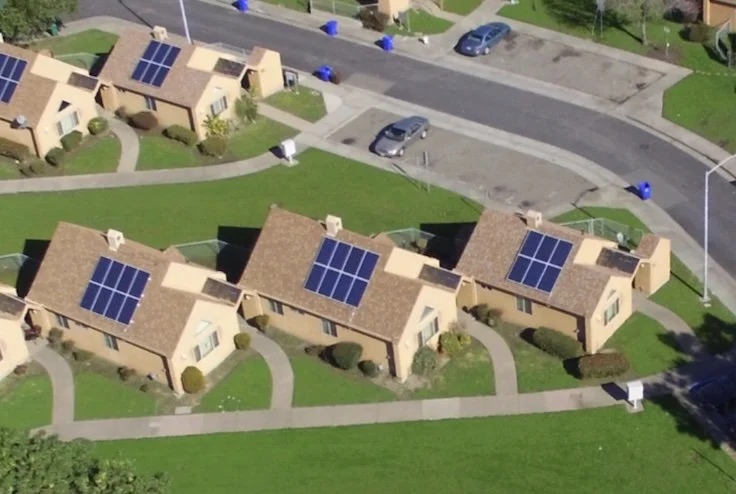 Aerial view of suburban houses with solar panels on brown roofs, surrounded by green lawns and walkways next to a paved road.