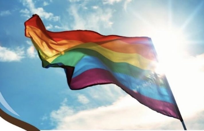 A six-striped rainbow pride flag waves dynamically against a bright blue sky with light clouds and a strong sun flare.