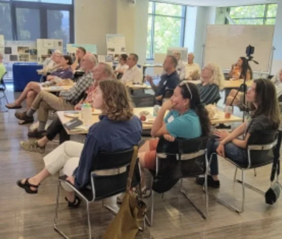 An audience sits at tables in a brightly lit classroom or conference room, looking toward a presentation off-camera.