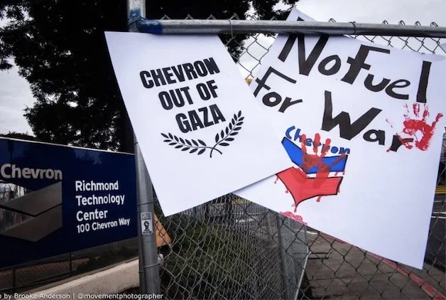 Protest signs reading "Chevron Out of Gaza" and "No Fuel for War" are attached to a chain-link fence at the Chevron Richmond Technology Center.