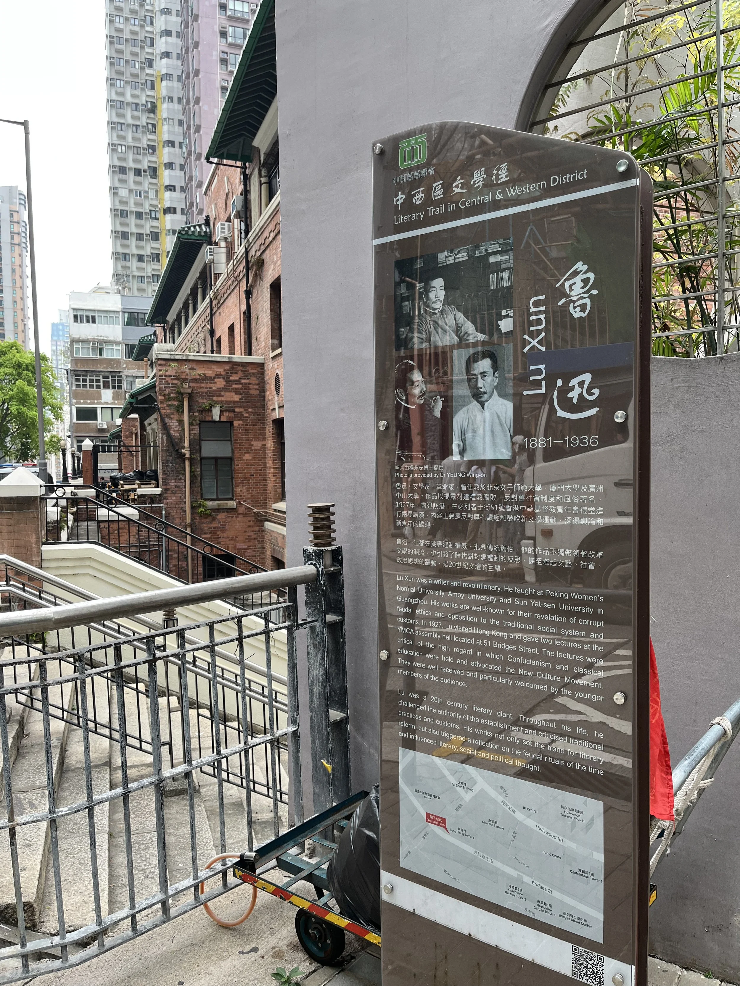 Information board about Lu Xun, a writer and revolutionary, located at a historical site in an urban area with high-rise buildings and a metal railing in the foreground.