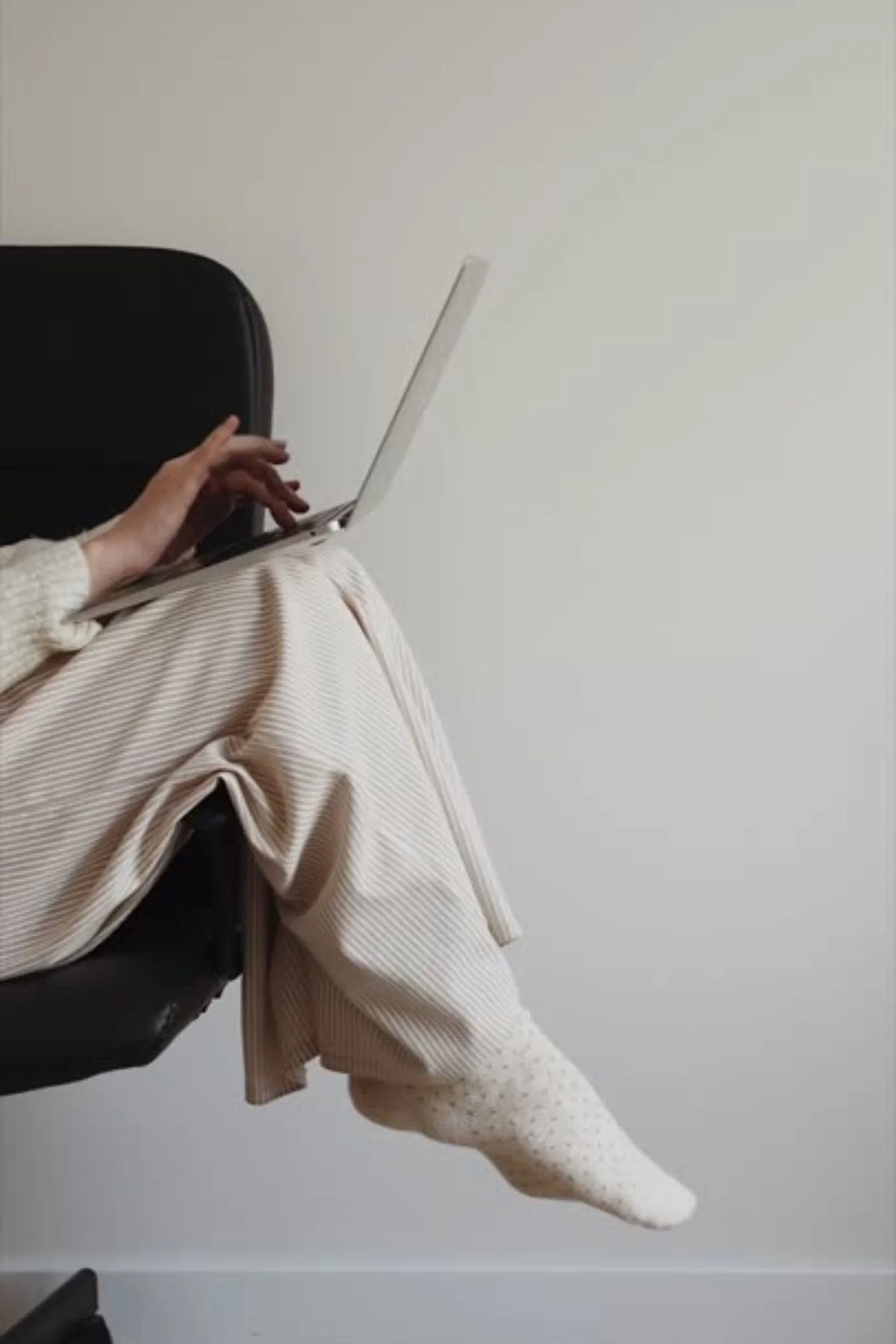 Person sitting on a chair using a laptop, with one leg extended and wearing a sock and beige pants, against a plain white wall.