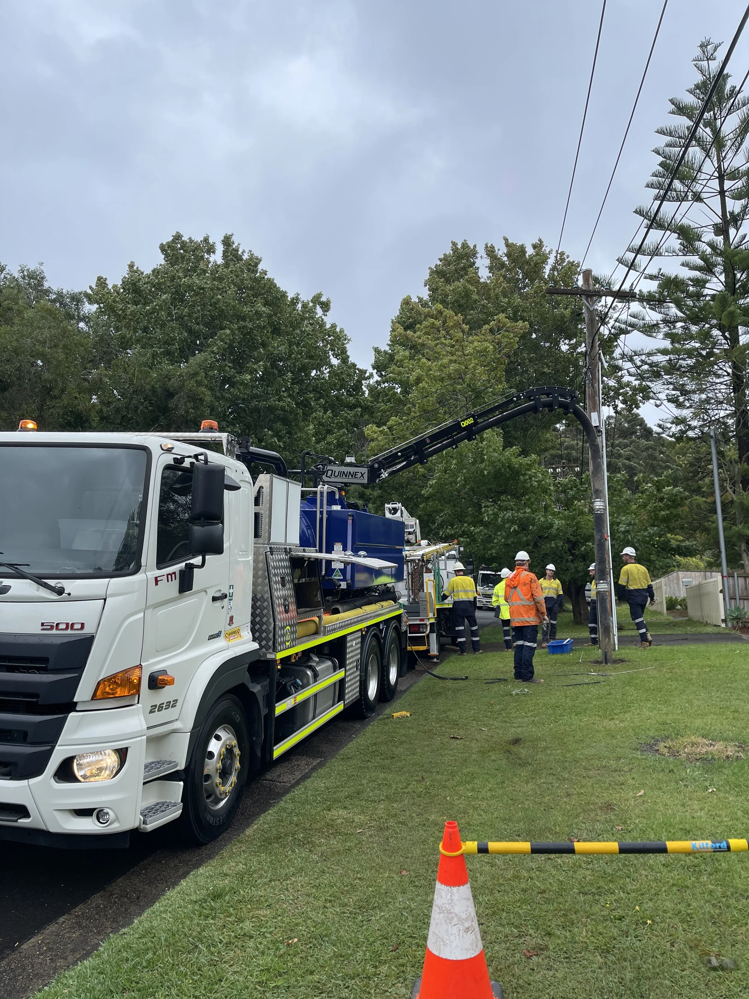Workers in safety gear fixing electrical lines on utility pole using a bucket truck on a residential street with trees and a grass lawn, with traffic cones and barrier.