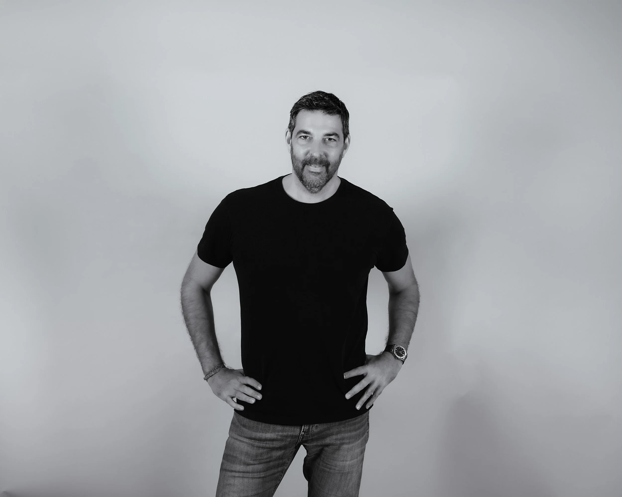 A man with dark hair and a beard posing with his hands on his hips, wearing a black t-shirt and jeans, against a plain light-colored background.
