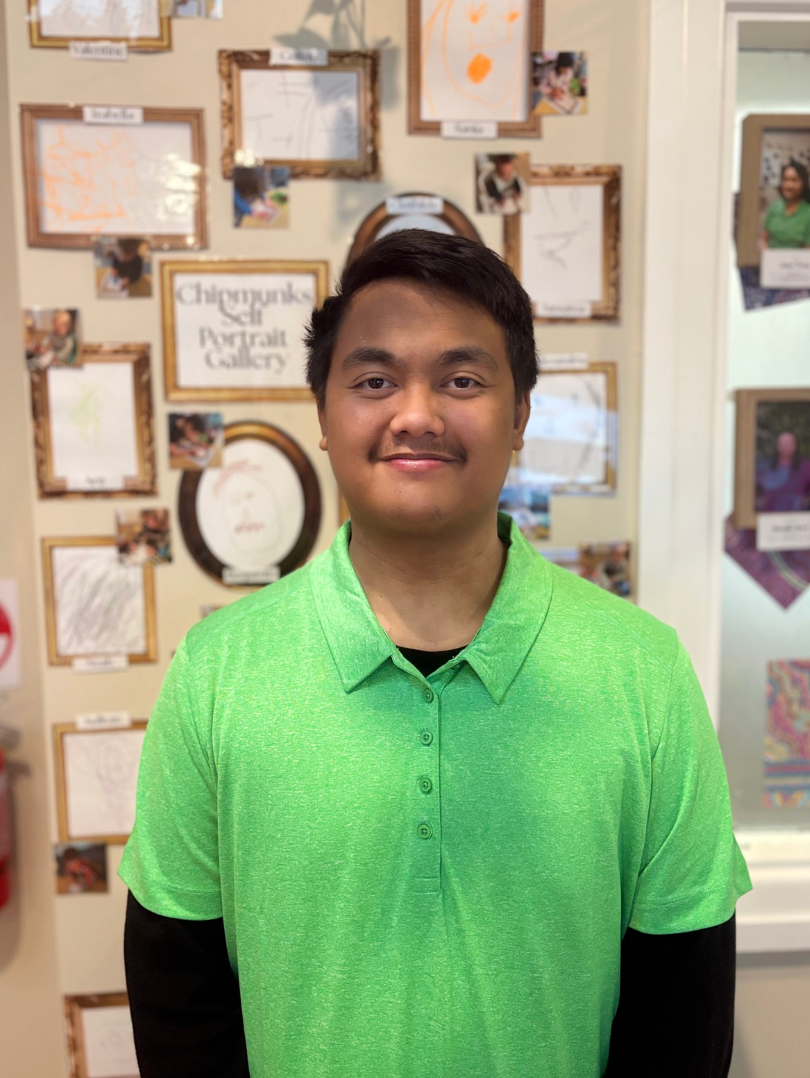 A young man with short black hair and a slight smile, wearing a bright green polo shirt over a black long-sleeve shirt, standing in front of a wall decorated with numerous picture frames and children's drawings.