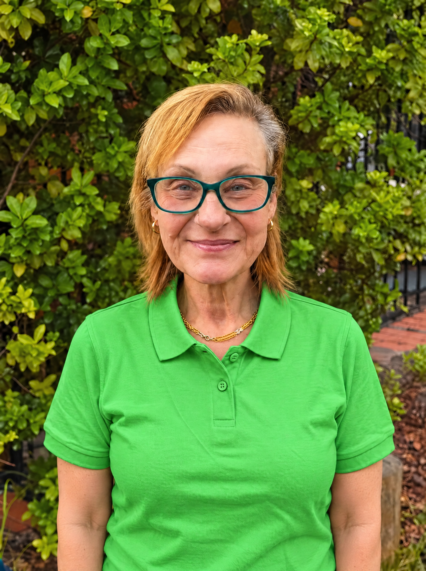 A woman with light skin, red hair, wearing green glasses, a black vest, green shirt, and gold necklace standing in front of a brick wall.