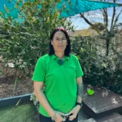 Person wearing a green shirt stands outdoors with trees and a blue canopy in the background.