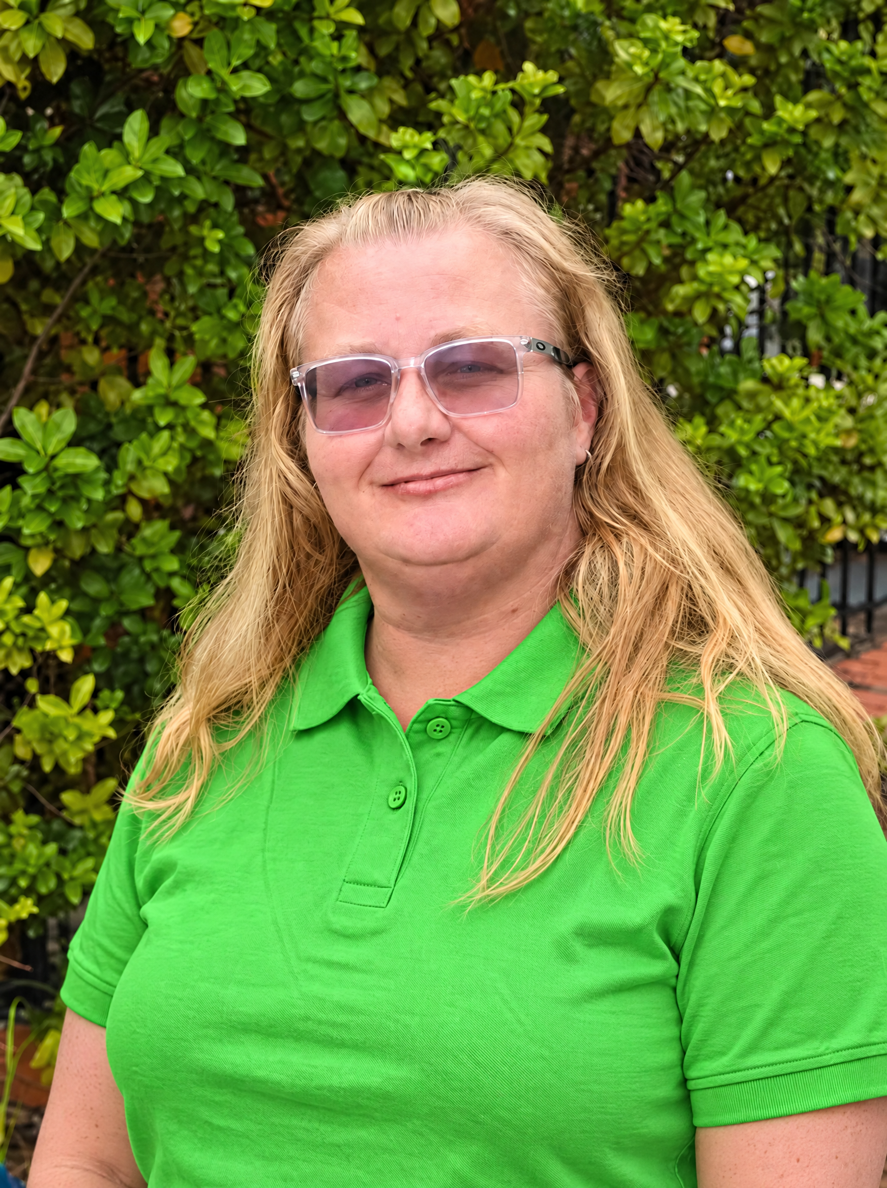A woman with long blonde hair and glasses, wearing a black hoodie, standing outdoors in front of greenery.