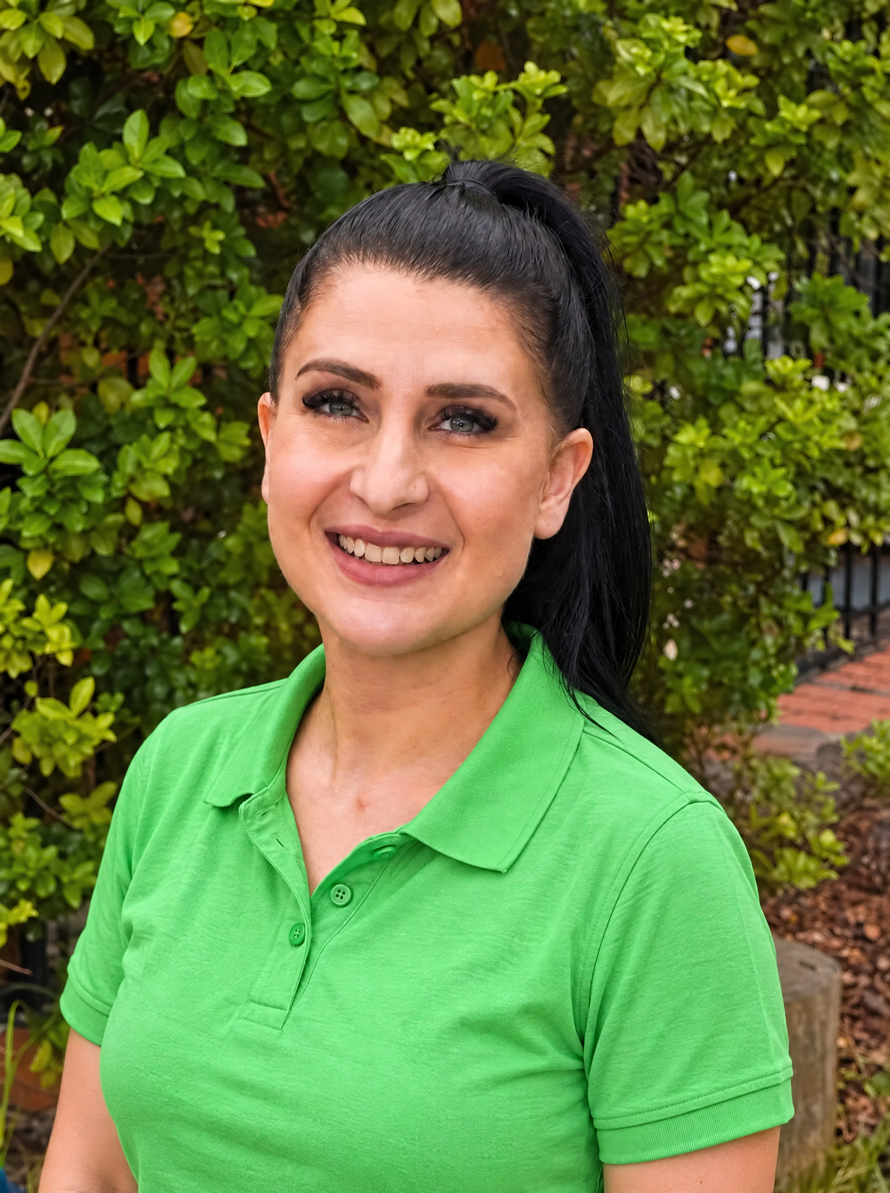 A young woman with dark hair in a ponytail, wearing a light green polo shirt, smiling outdoors in front of green foliage.