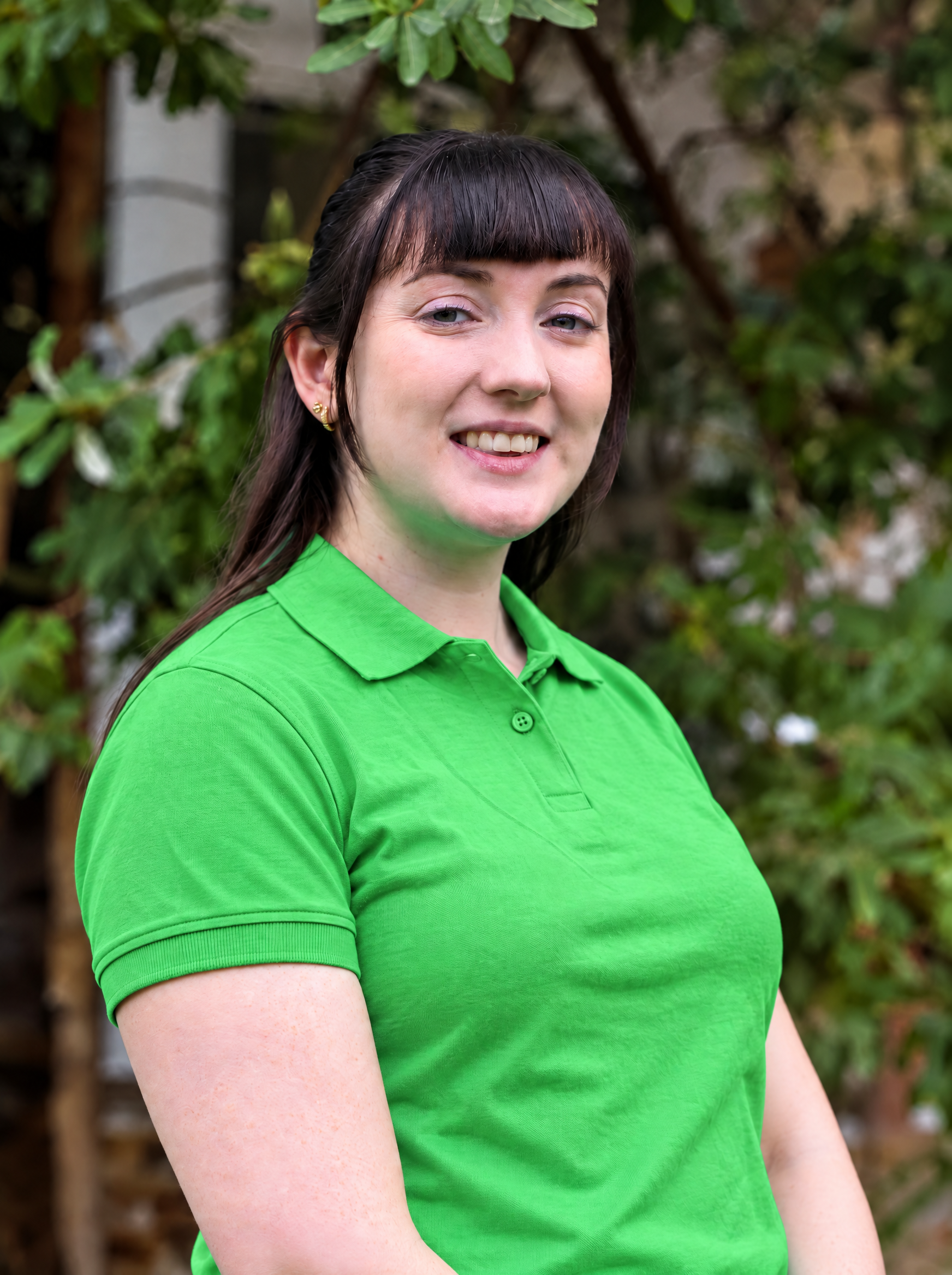 Young woman with dark hair, wearing a bright green polo shirt, standing indoors with a blurred background of plants and decorations.