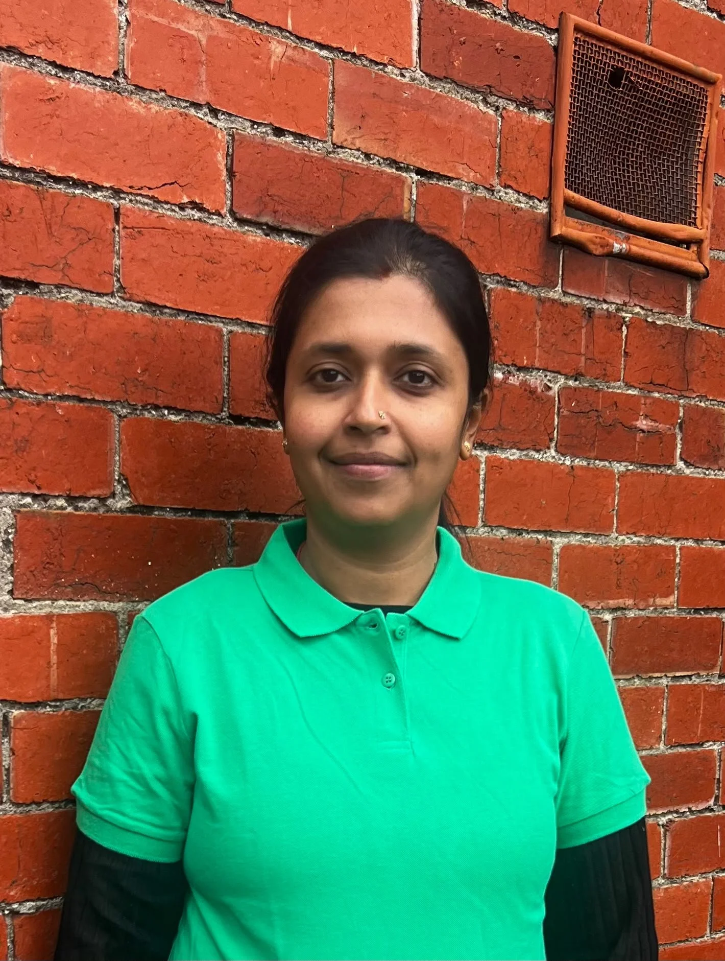 A woman with blonde hair tied back, wearing a green polo shirt and a black Adidas jacket, standing outdoors in front of green foliage.