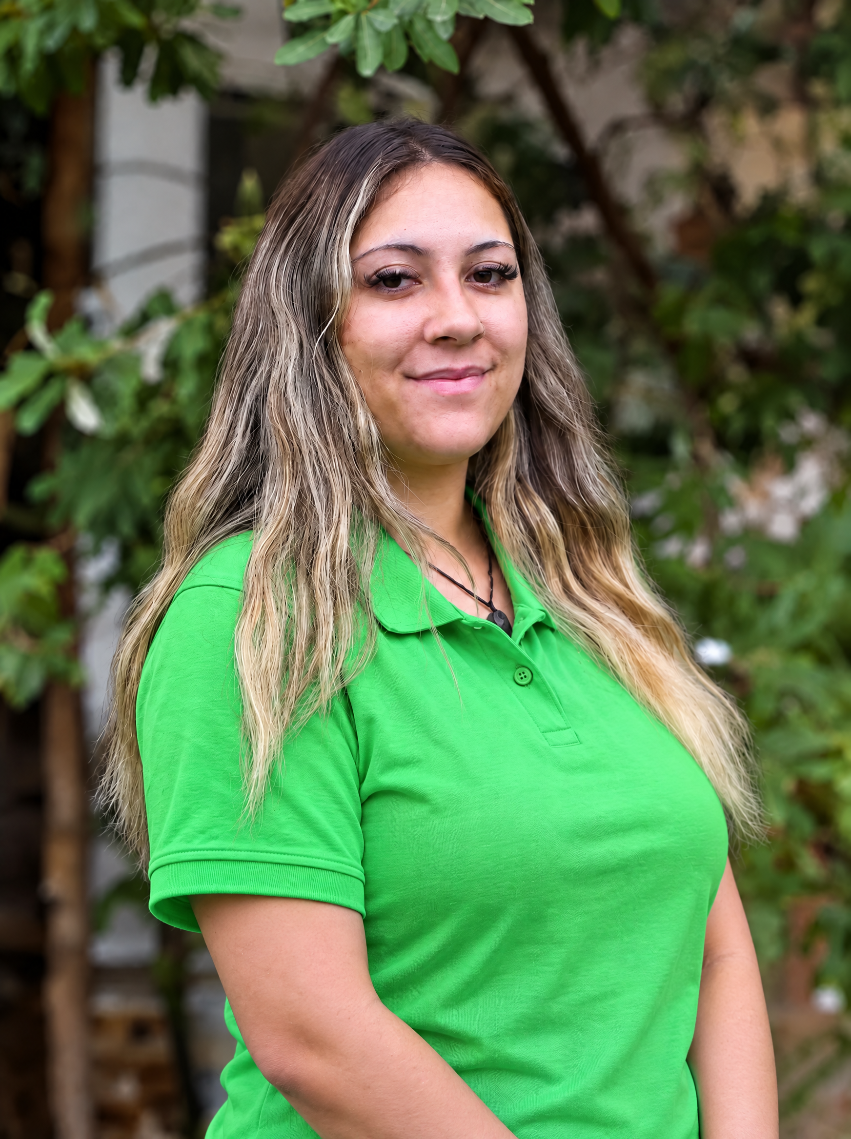 Person with long hair and glasses wearing a green shirt, standing outdoors near a tree.
